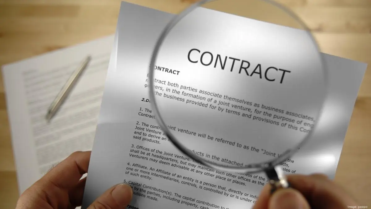 Hand holding a magnifying glass over a document titled 'CONTRACT' with a pen nearby.